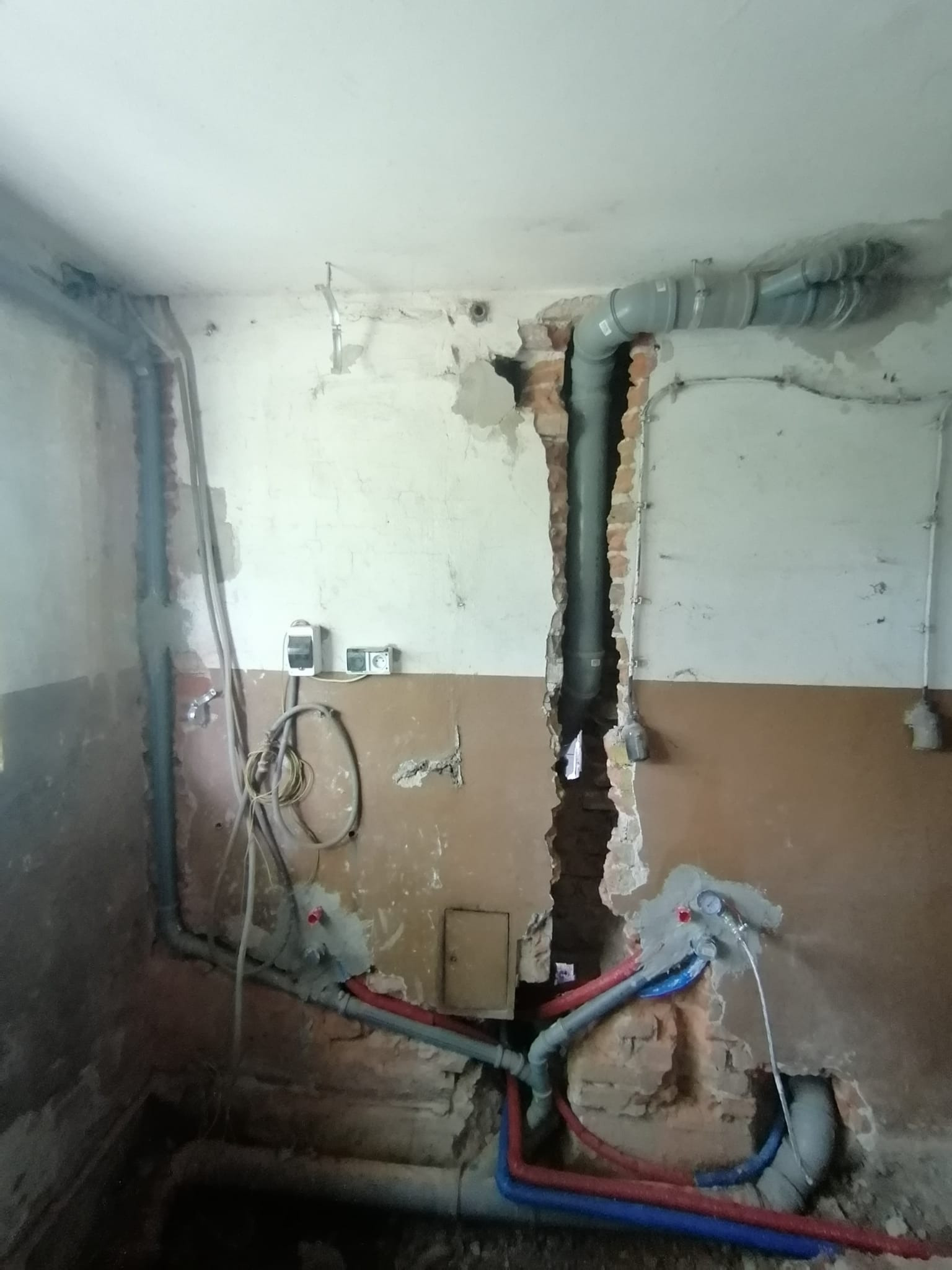Odsłonięte rury kanalizacyjne i wodne w trakcie prac hydraulicznych w pomieszczeniu z częściowo otynkowanymi ścianami, widoczne fragmenty cegieł i instalacji elektrycznej.