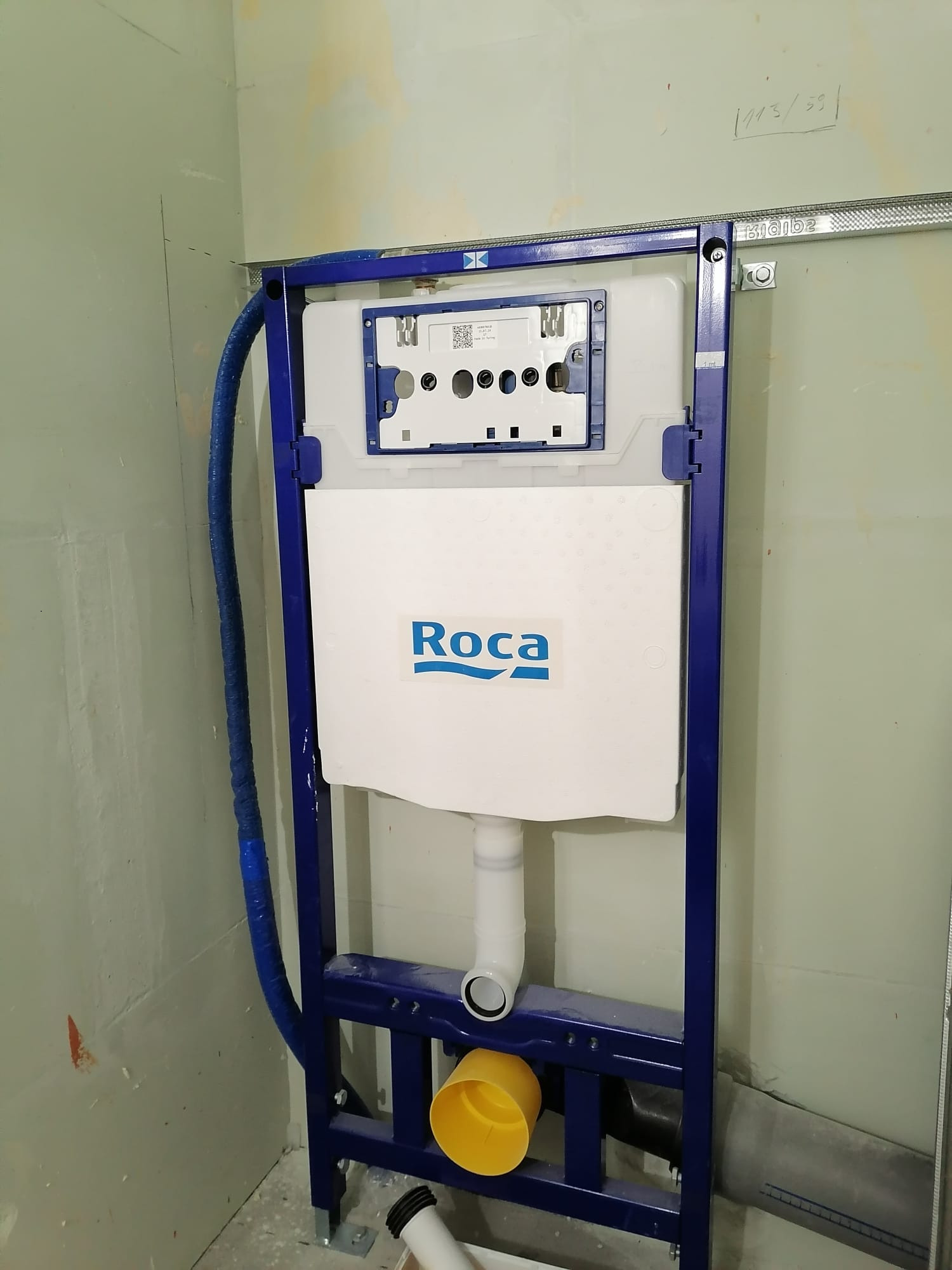 Stelaż podtynkowy WC marki Roca, zamontowany na tle surowej ściany, z widocznymi elementami instalacji hydraulicznymi i odpływem kanalizacyjnym.