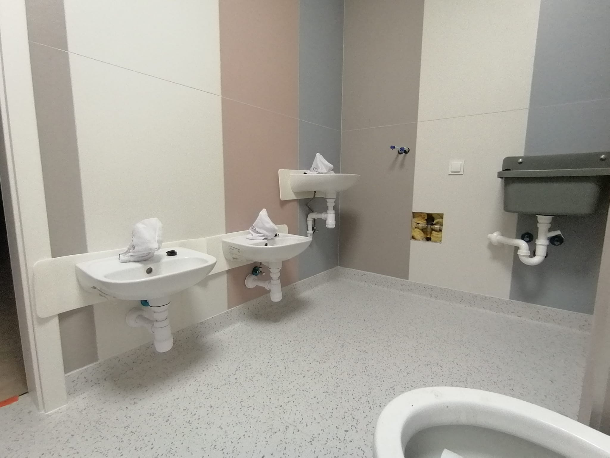 Wnętrze łazienki w szpitalu lub placówce medycznej z trzema umywalkami na ścianie w kolorowe pasy, widoczny fragment toalety.