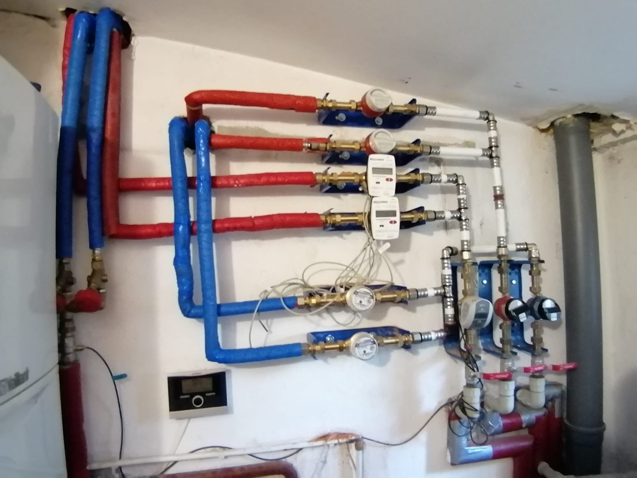Rozbudowana instalacja wodna i grzewcza z rurami w izolacji termicznej (czerwonej i niebieskiej), liczniki wody, zawory, czujniki oraz panel sterowania na ścianie w pomieszczeniu technicznym.