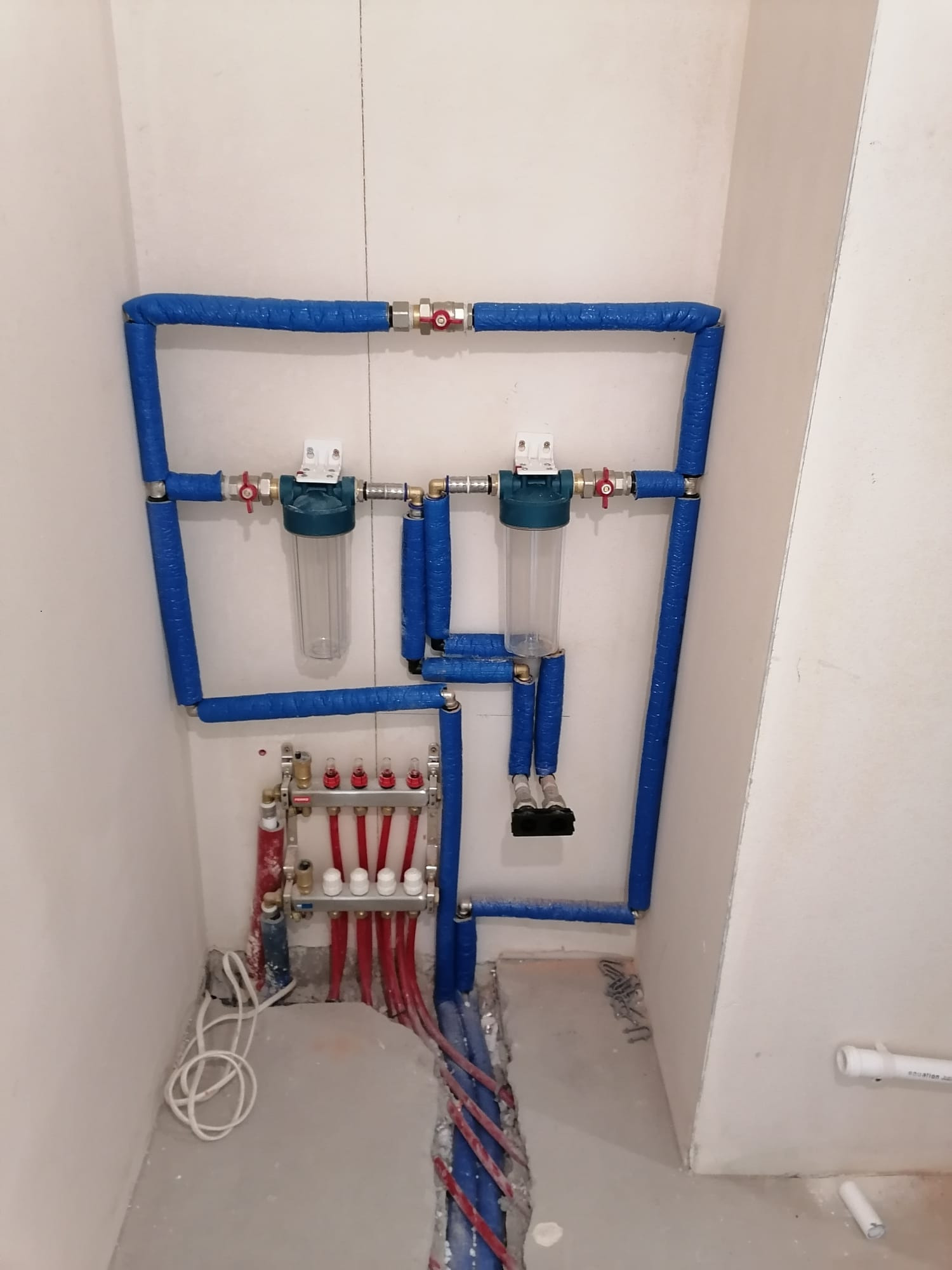 Instalacja hydrauliczna z filtrami wody i rozdzielaczem ogrzewania podłogowego, rury w niebieskiej izolacji, widok na surowy stan podłogi.