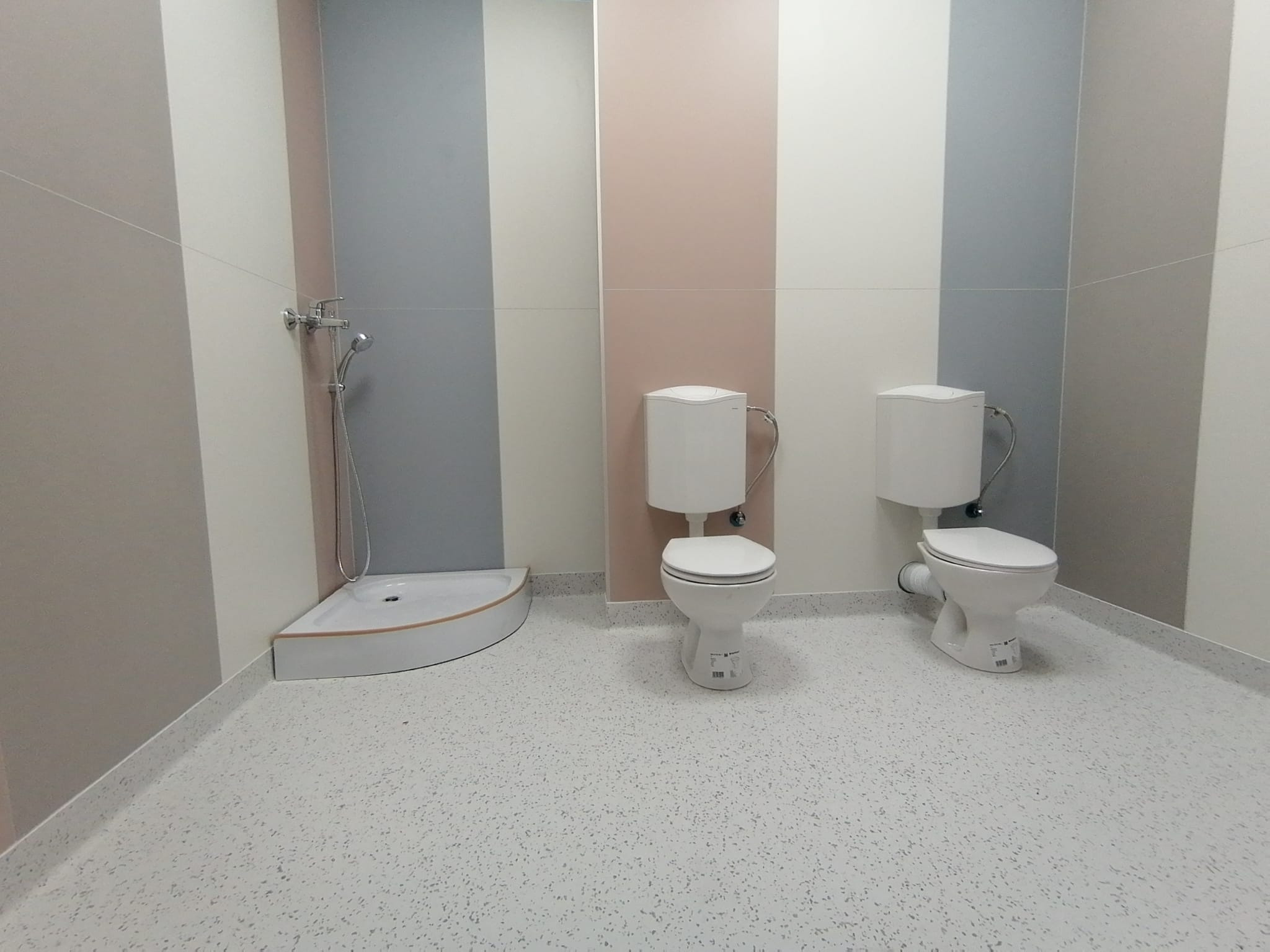 Jasna łazienka z dwoma toaletami i narożnym brodzikiem, ściany w pionowe pasy beżu, szarości i różu, podłoga w jasne plamki.