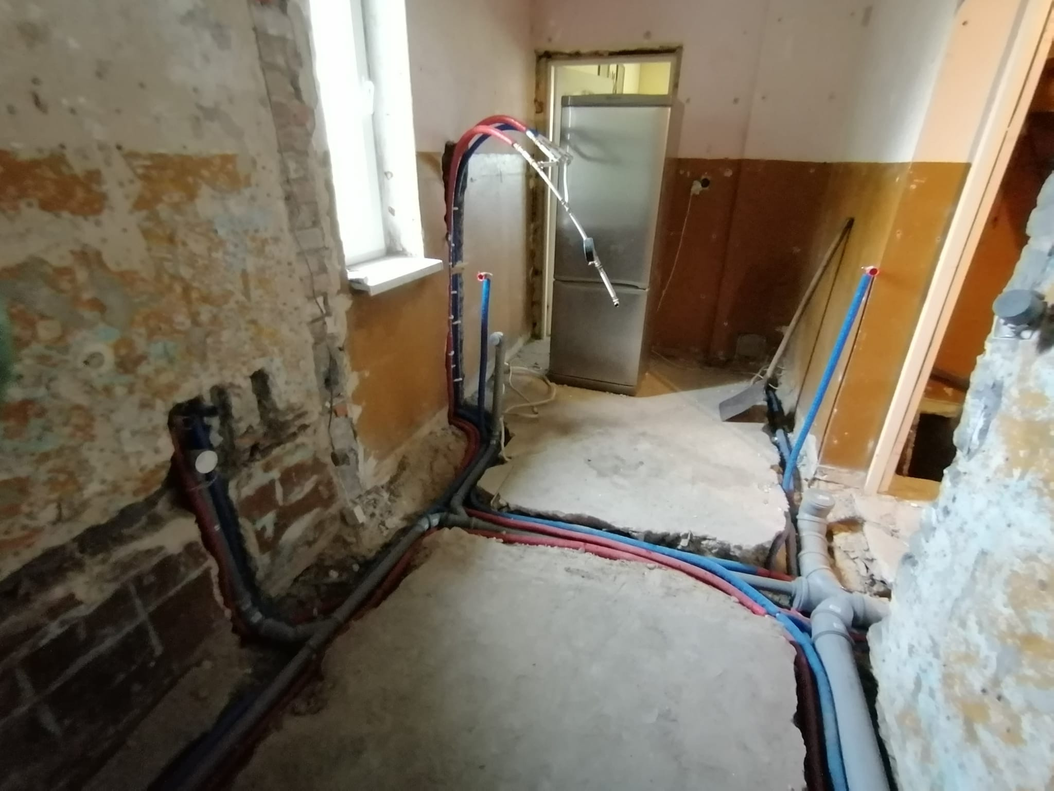 Wnętrze w trakcie remontu z odsłoniętymi instalacjami hydraulicznymi; widoczne rury w kolorach czerwonym i niebieskim prowadzone w bruzdach w podłodze i ścianach, surowe ściany z widoczną cegłą,...