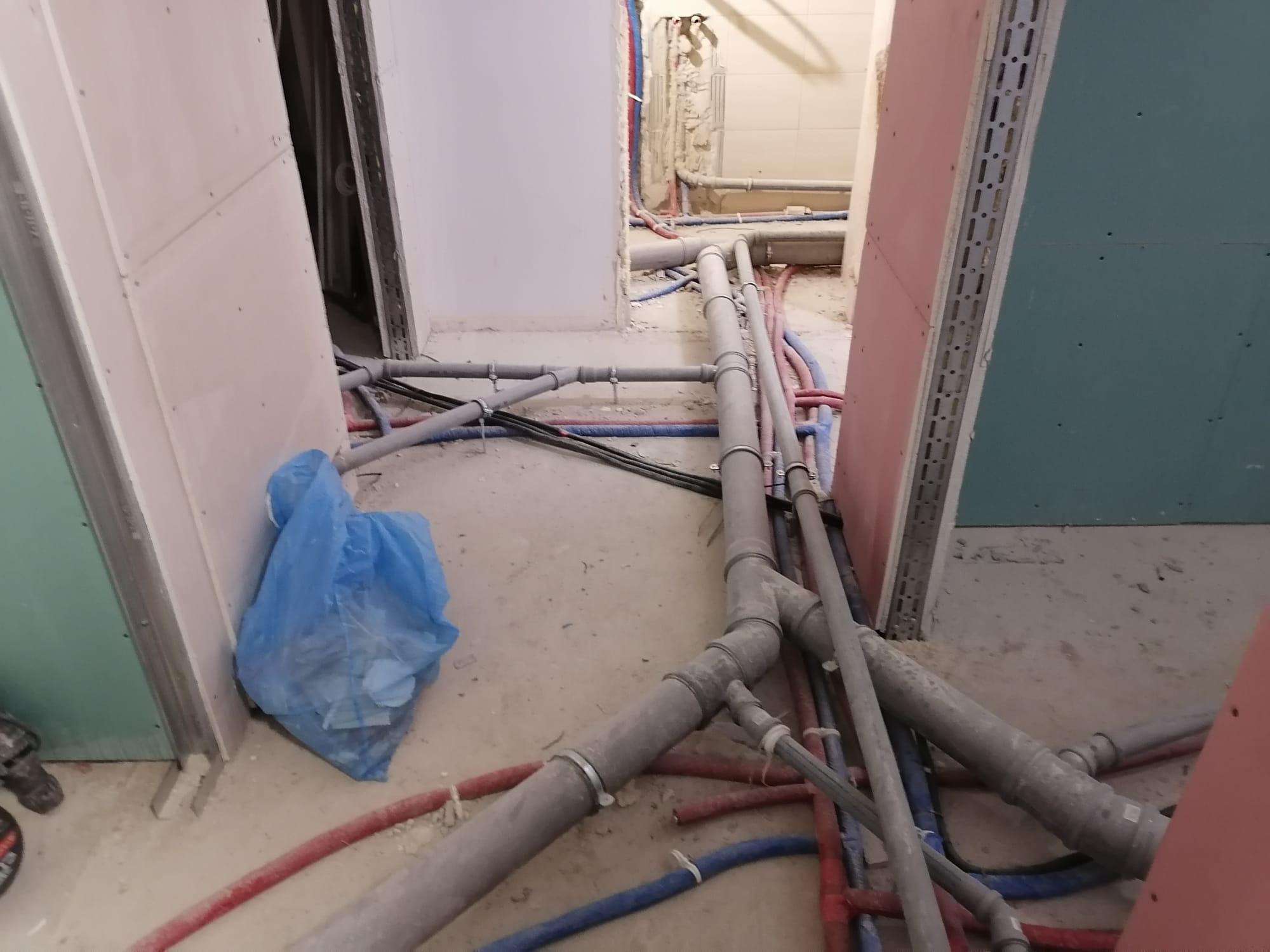 Wnętrze w trakcie remontu z widocznymi rurami instalacji hydraulicznej i grzewczej, częściowo zamontowanymi, podłoga pokryta kurzem i resztkami materiałów budowlanych, ściany w surowym stanie...