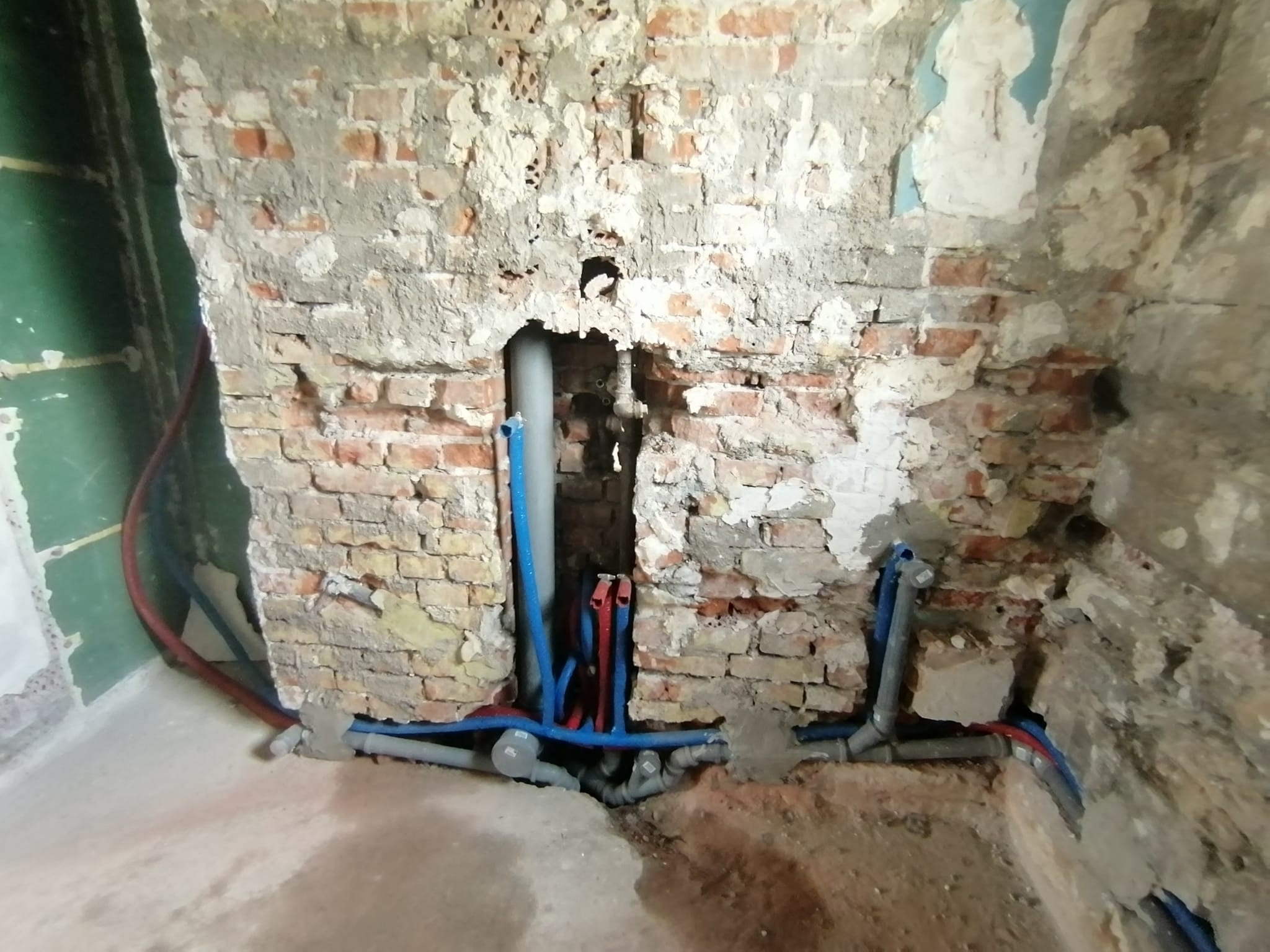 Surowa ściana z cegły z widocznymi nowymi instalacjami hydraulicznymi: niebieskie i czerwone rury prowadzone w bruzdach, szare rury odpływowe na podłodze, ślady po kuciu w ścianie.