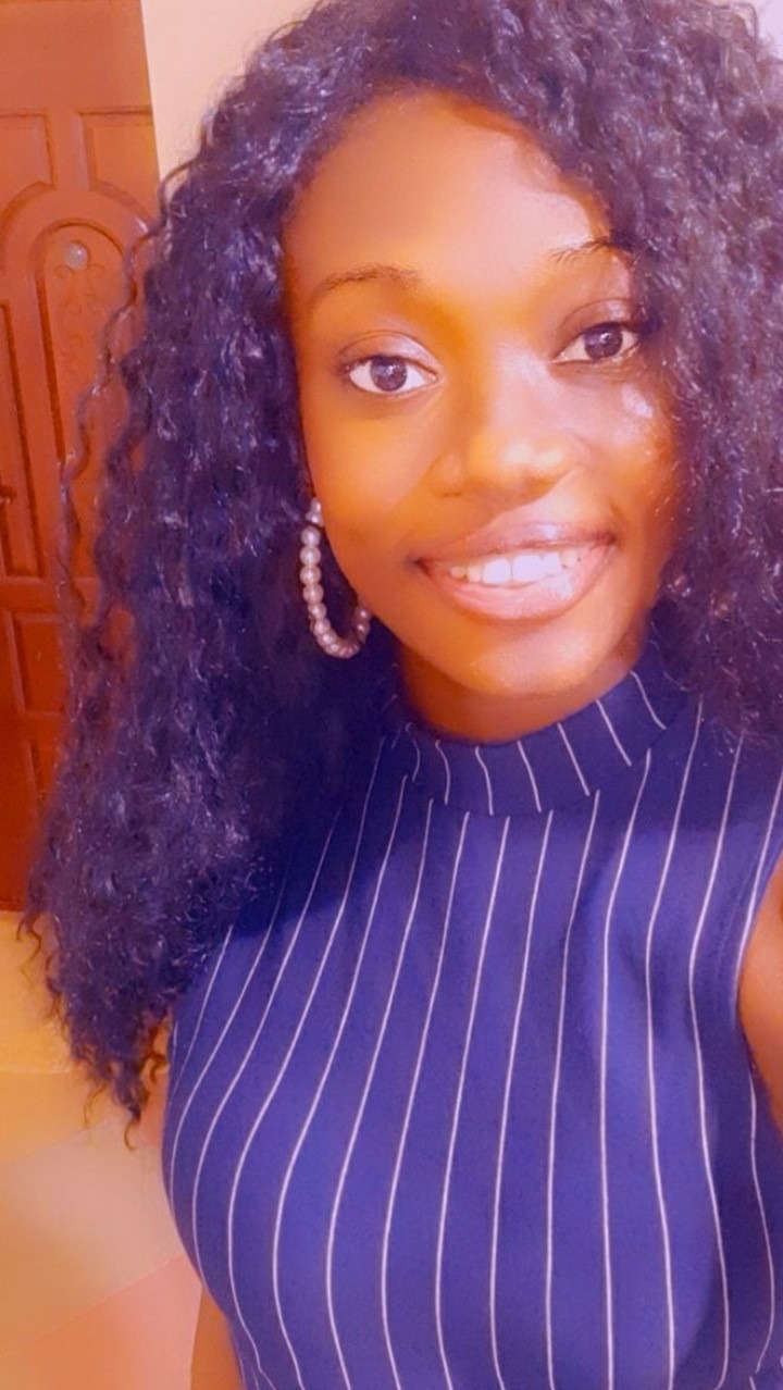 Portret uśmiechniętej kobiety z kręconymi włosami, w bluzce w paski i perłowych kolczykach, pozującej na tle drewnianych drzwi.