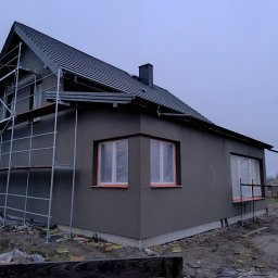 Remonty z pasją Mariusz Dopierała - Dom jednorodzinny w trakcie tynkowania elewacji zewnętrznej, widoczne rusztowania i zabezpieczone okna folią malarską.