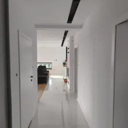 Nowoczesny korytarz z białymi ścianami, błyszczącą podłogą z płytek, czarnymi listwami sufitowymi z oświetleniem punktowym oraz widokiem na salon z brązową sofą i oknem.