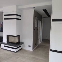 Remonty z pasją Mariusz Dopierała - Nowoczesny salon po remoncie: białe ściany z czarnymi akcentami, kominek z czarnym blatem, jasna podłoga z płytek imitujących drewno, czarny worek na śmieci w rogu.