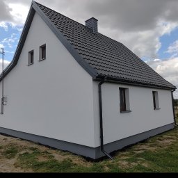 Remonty z pasją Mariusz Dopierała - Boczny widok domu z białymi ścianami i szarym dachem. Widoczne okna, rynna i fragment ogrodu. Dom po świeżym malowaniu elewacji i podmurówki.