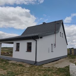 Remonty z pasją Mariusz Dopierała - Biały dom jednorodzinny z szarą dachówką i podmurówką, widok z boku w słoneczny dzień. Widoczne okna z brązowymi ramami i elementy wykończeniowe.