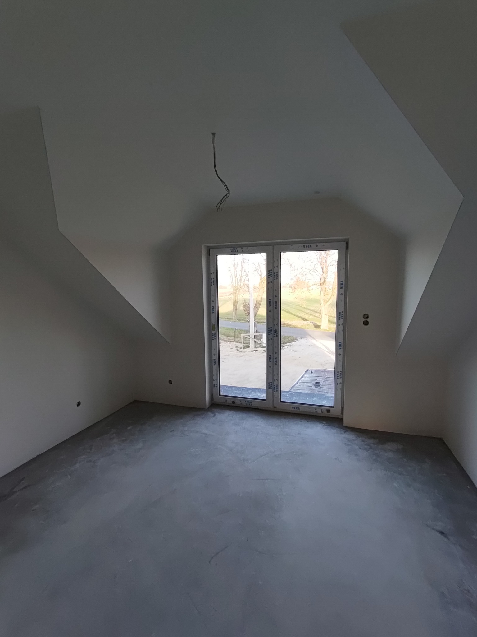 Surowe wnętrze poddasza z oknem balkonowym, widok na zewnątrz, ściany przygotowane pod gładzie, wylewka podłogowa w szarym kolorze, wystające przewody elektryczne.
