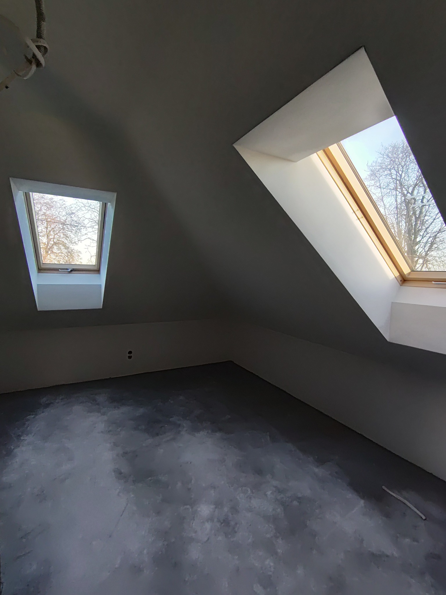 Poddasze po nałożeniu gładzi, widoczne dwa okna dachowe z drewnianymi ramami i widokiem na drzewa, ściany szare, podłoga pokryta warstwą wyrównującą.