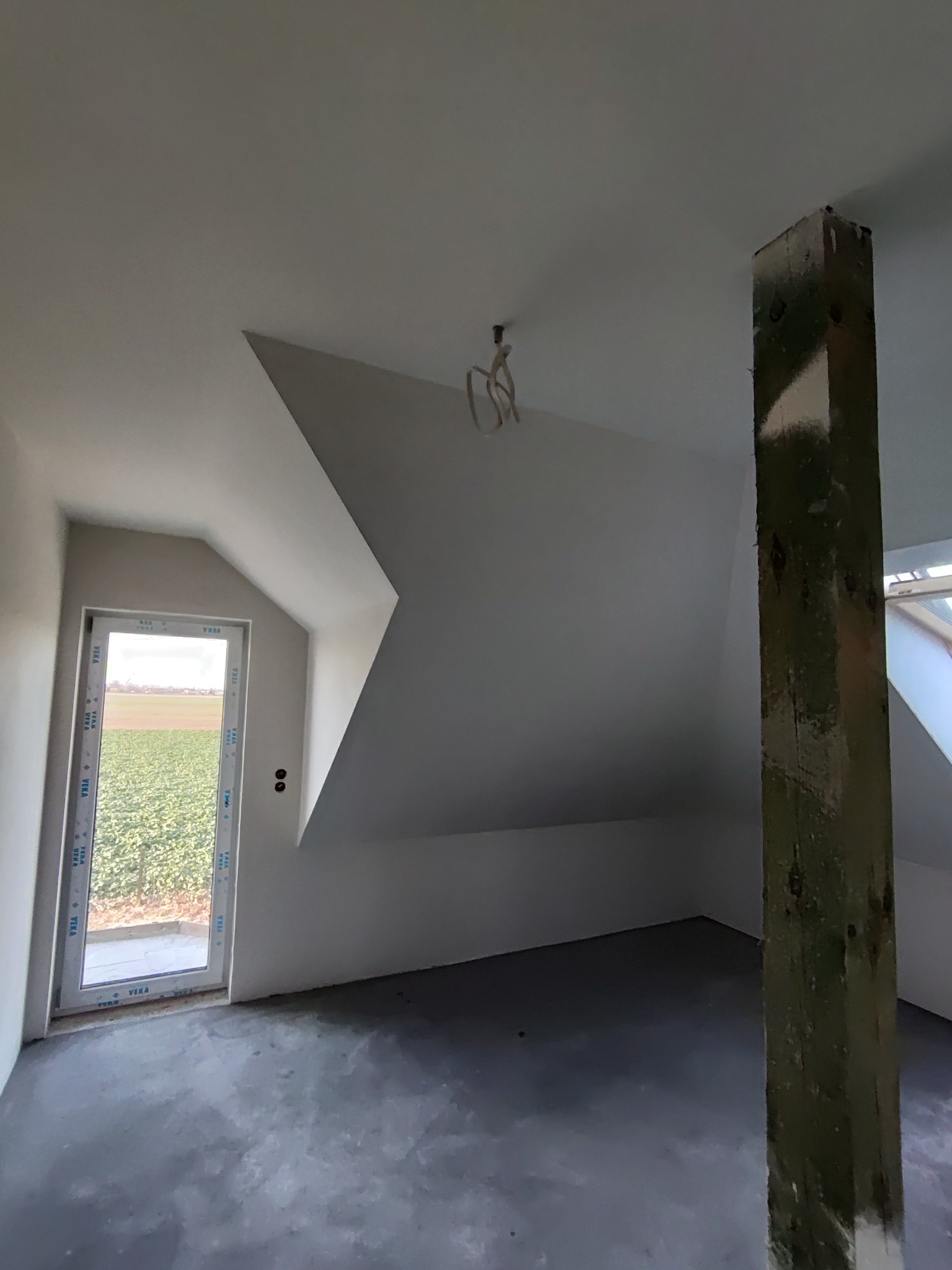 Surowe wnętrze poddasza z oknem dachowym i nieotynkowanym słupem nośnym, widoczne kable oświetleniowe i wylewka na podłodze.