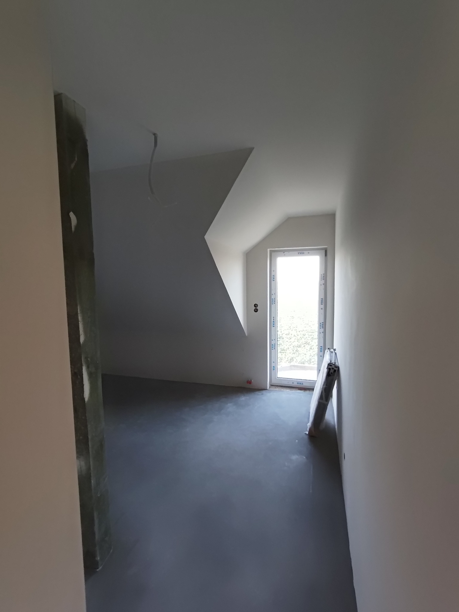 Poddasze w trakcie remontu: gładzie na ścianach, wylewka na podłodze, okno dachowe z widokiem na zieleń, widoczne przewody elektryczne.