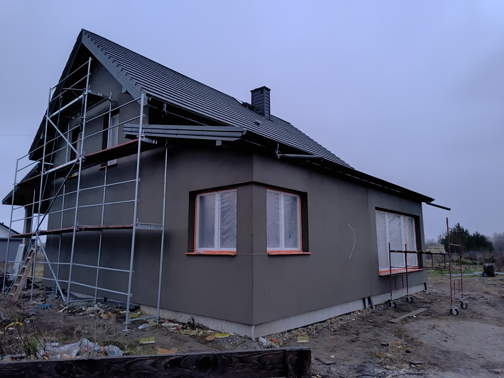 Dom jednorodzinny w trakcie tynkowania elewacji zewnętrznej, widoczne rusztowania i zabezpieczone okna folią malarską.