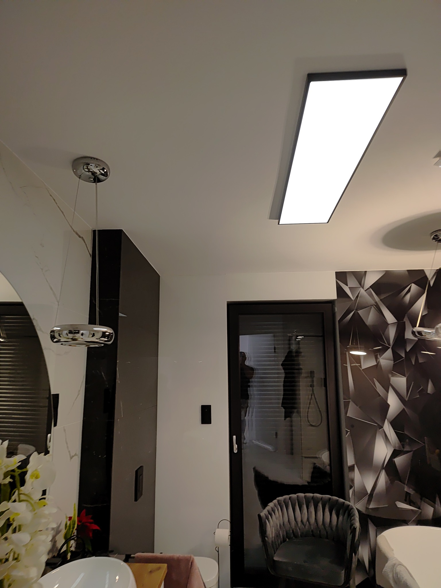 Nowoczesna łazienka z podwieszaną lampą LED, chromowaną armaturą, umywalką nablatową i geometryczną tapetą akcentującą ścianę.