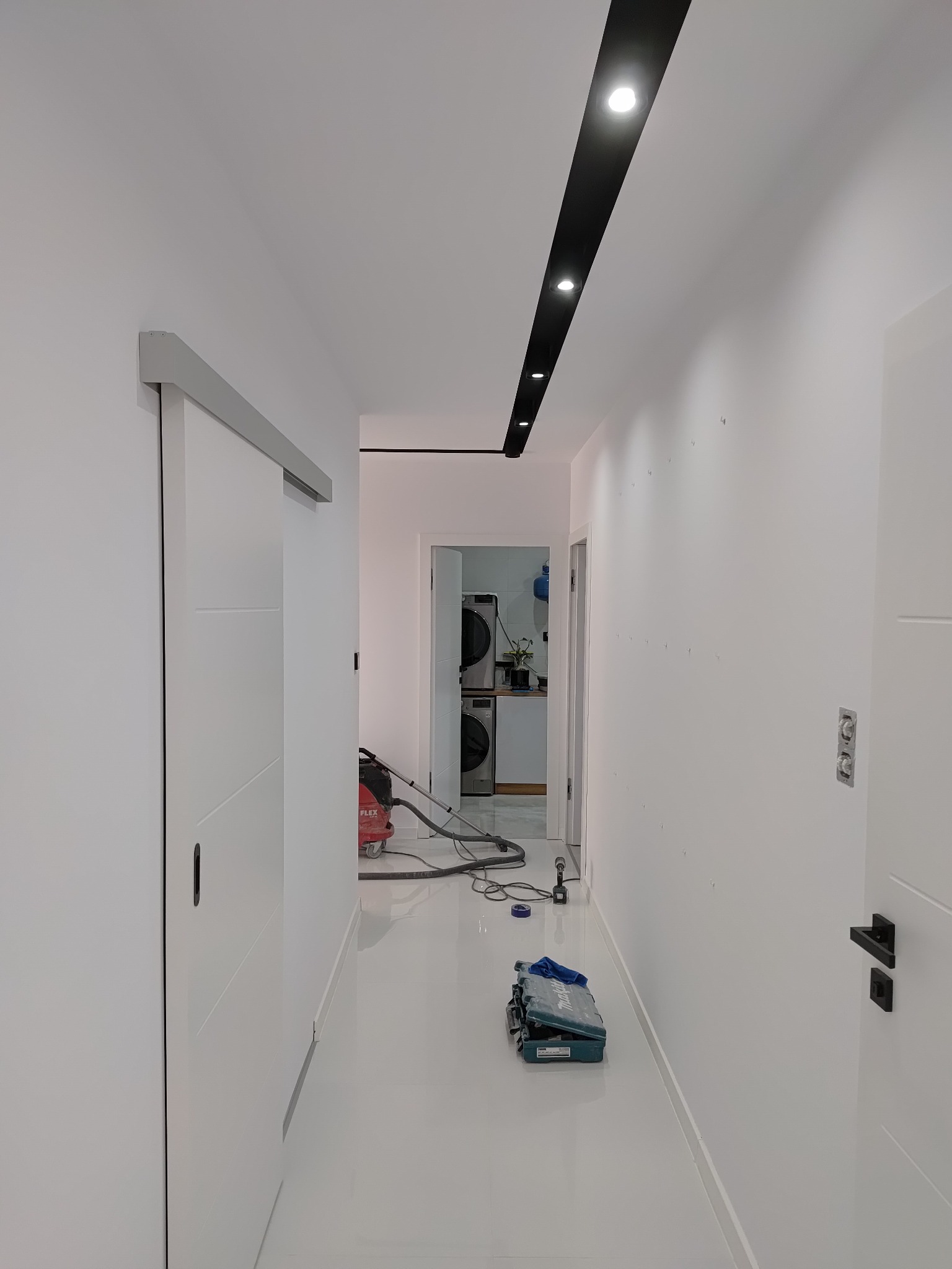 Biały korytarz po remoncie z nowymi przesuwnymi drzwiami, oświetleniem sufitowym w czarnej listwie i błyszczącą podłogą, widoczny odkurzacz i skrzynka z narzędziami, ślady po szpachlowaniu na ścianie.