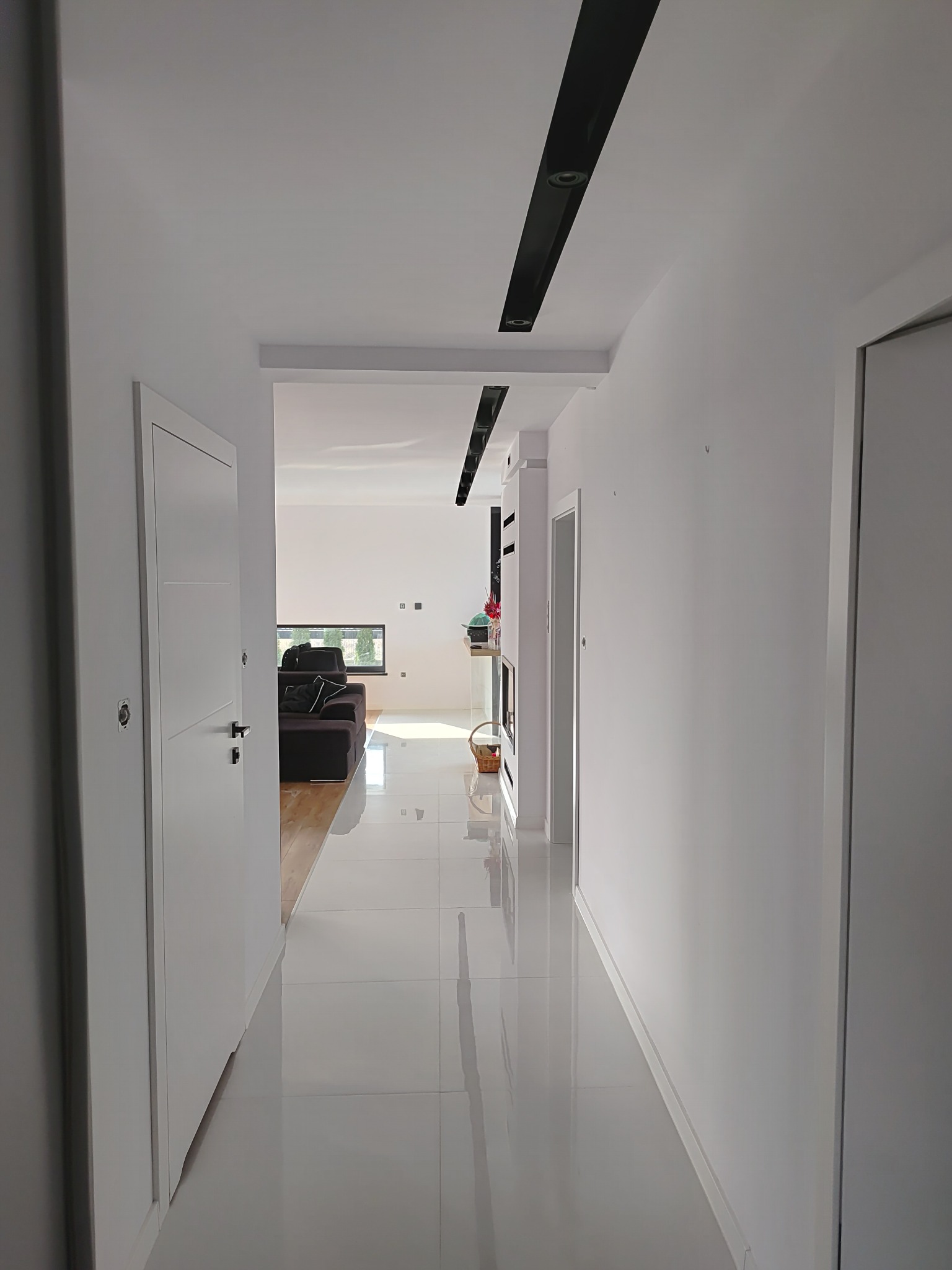 Nowoczesny korytarz z białymi ścianami, błyszczącą podłogą z płytek, czarnymi listwami sufitowymi z oświetleniem punktowym oraz widokiem na salon z brązową sofą i oknem.