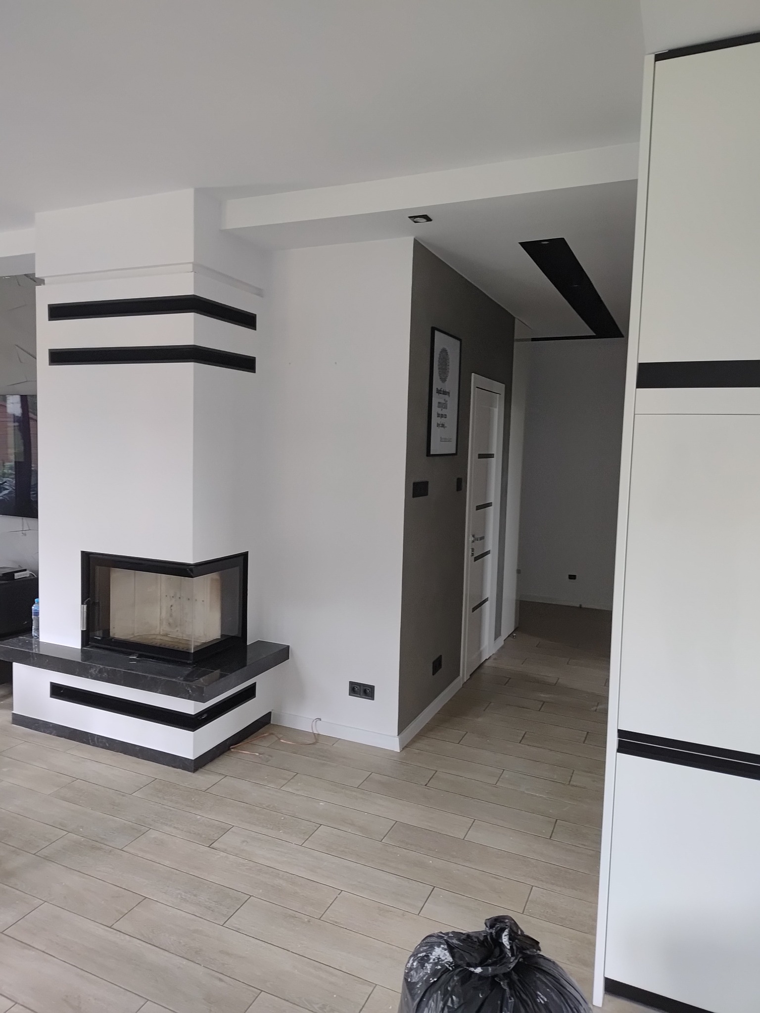 Nowoczesny salon po remoncie: białe ściany z czarnymi akcentami, kominek z czarnym blatem, jasna podłoga z płytek imitujących drewno, czarny worek na śmieci w rogu.