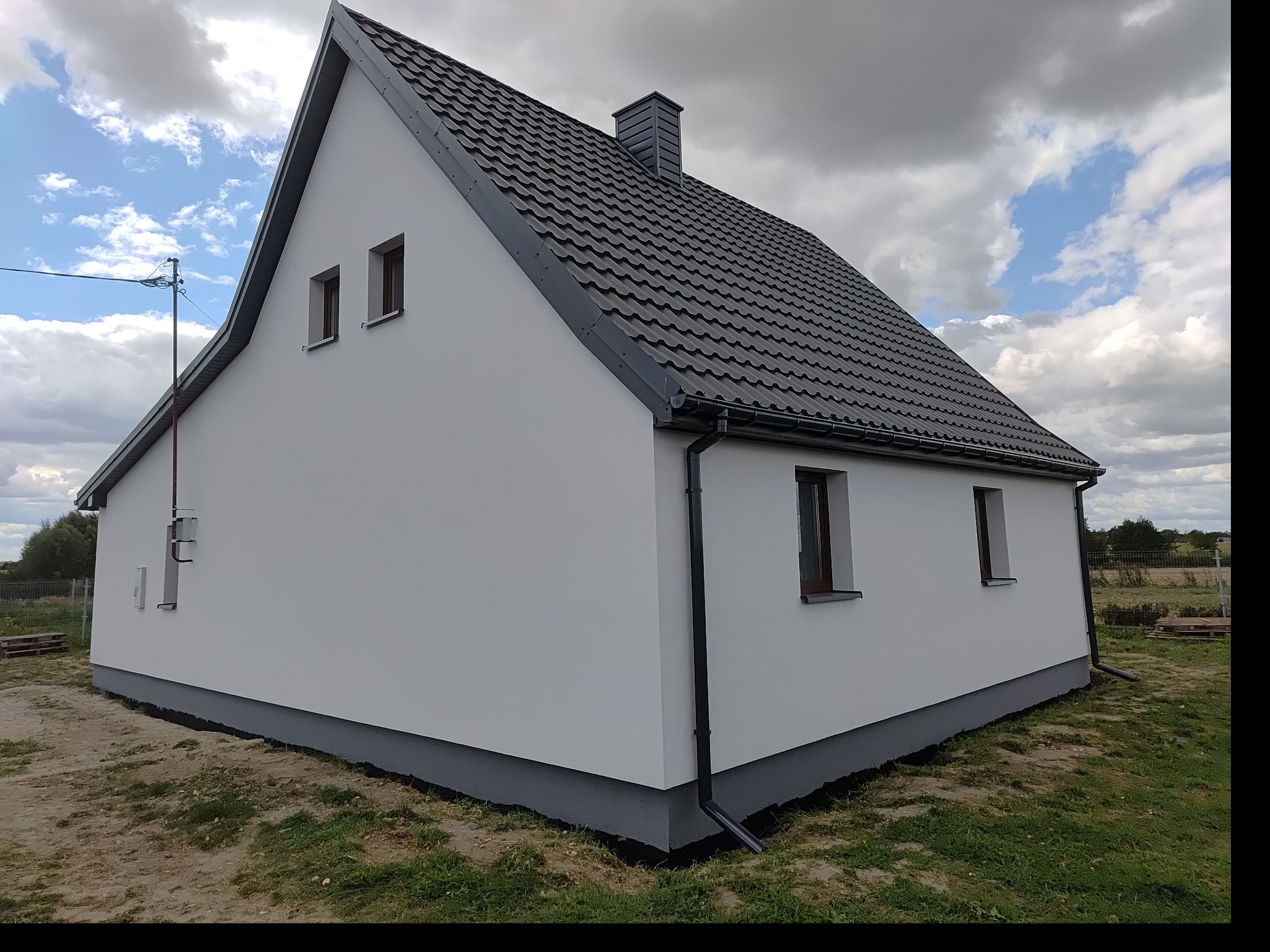 Boczny widok domu z białymi ścianami i szarym dachem. Widoczne okna, rynna i fragment ogrodu. Dom po świeżym malowaniu elewacji i podmurówki.