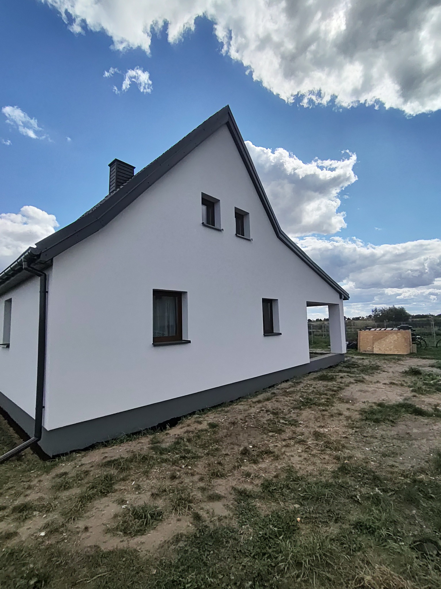 Biały dom jednorodzinny z szarym dachem i podmurówką na tle błękitnego nieba z chmurami. Widok z boku, ukazujący elewację po remoncie.