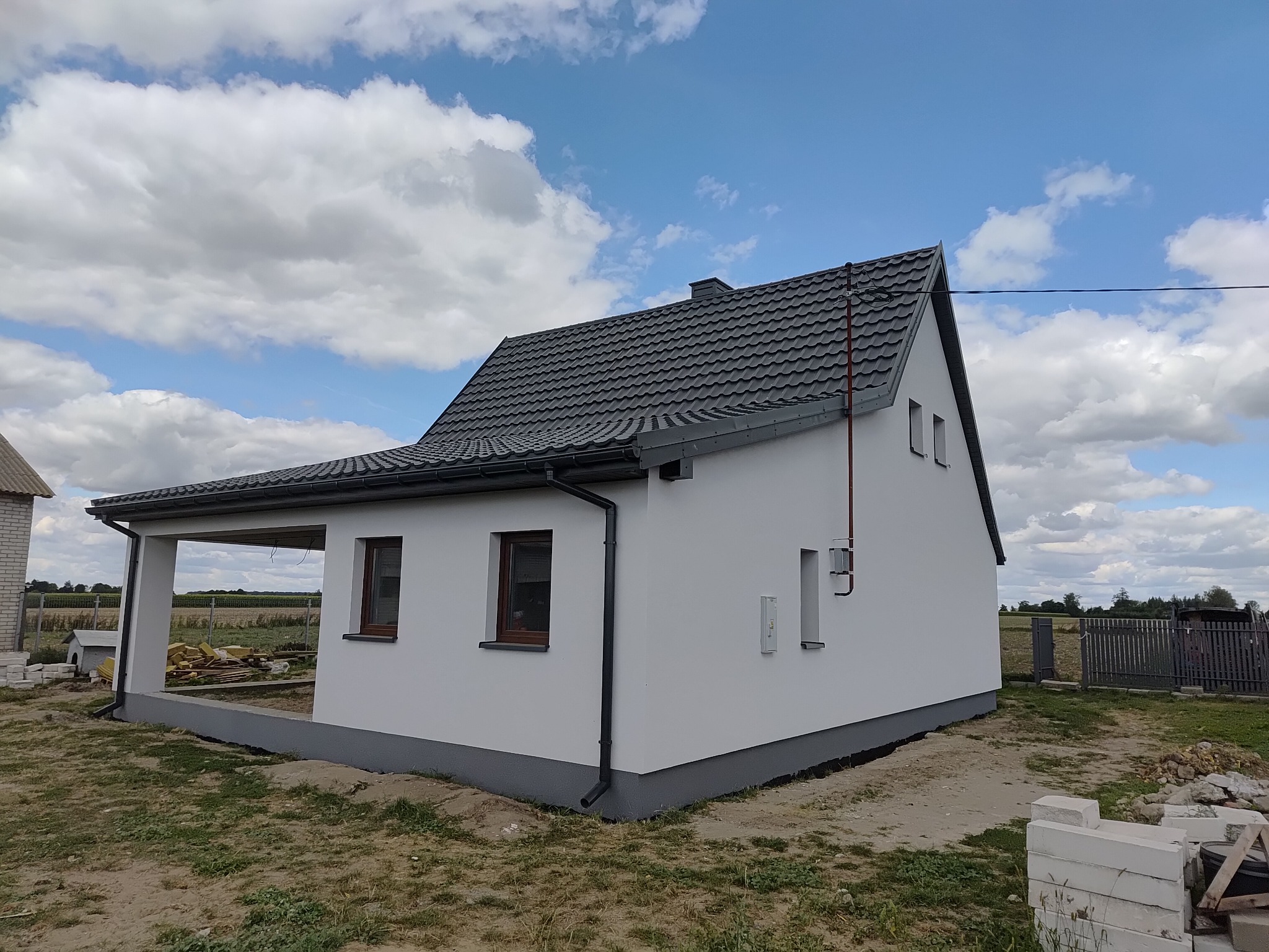 Biały dom jednorodzinny z szarą dachówką i podmurówką, widok z boku w słoneczny dzień. Widoczne okna z brązowymi ramami i elementy wykończeniowe.