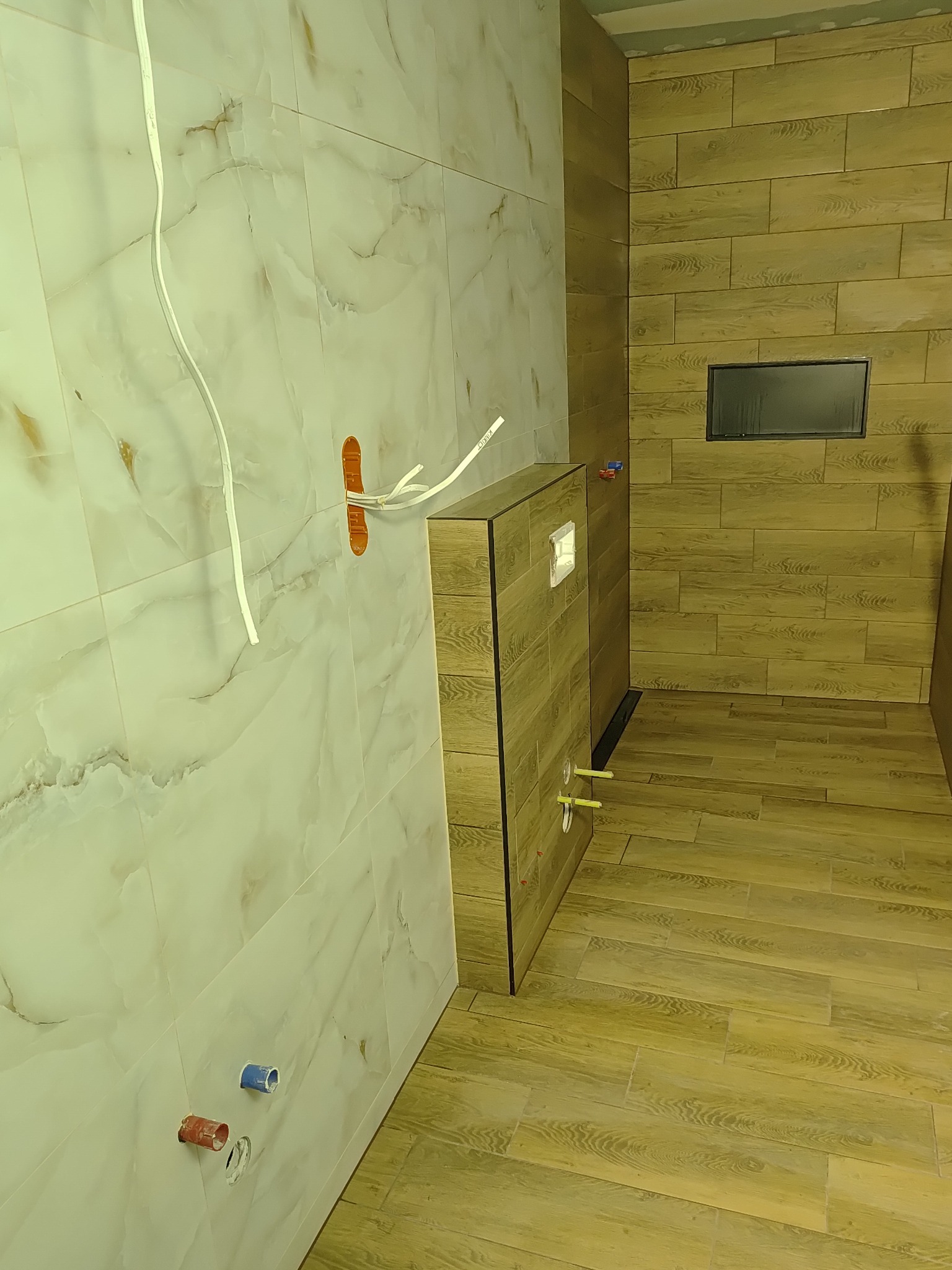 Łazienka w trakcie wykończenia: marmurowe płytki ścienne, drewnopodobne płytki na podłodze i ścianie, zabudowa WC z widocznymi przyłączami. Prace instalacyjne w toku.