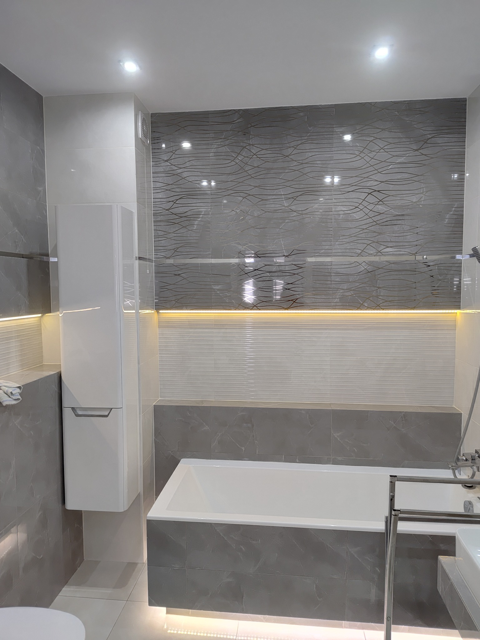 Nowoczesna łazienka w szarościach z podświetleniem LED. Uwagę przykuwają płytki imitujące marmur i dekoracyjne panele ścienne. Minimalistyczna szafka wisząca.