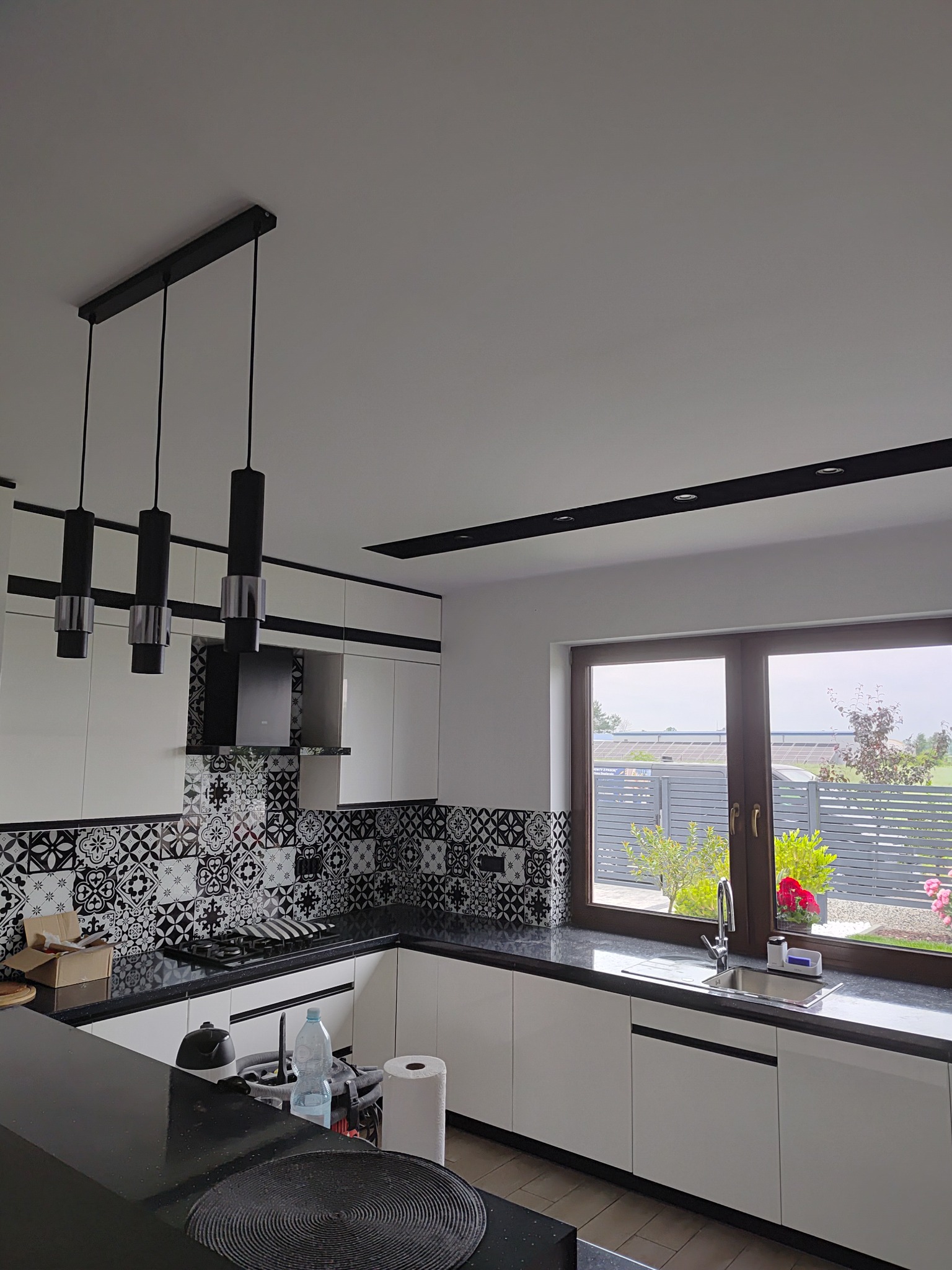 Nowoczesna kuchnia po remoncie: białe szafki, czarny blat, wzorzyste płytki nad blatem, okno z widokiem na ogród, designerskie lampy wiszące.