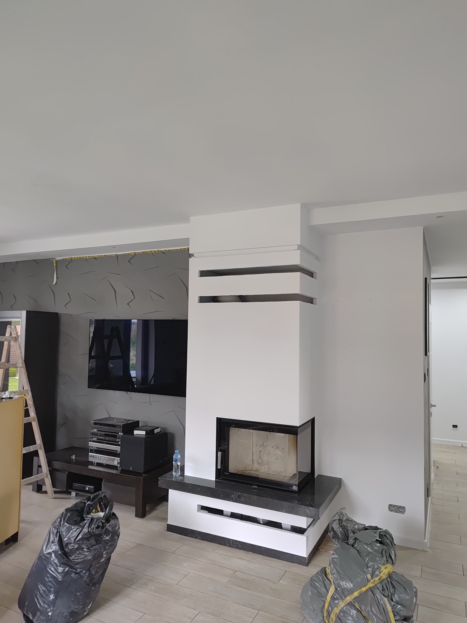 Nowoczesny kominek z czarnym obramowaniem i białymi elementami w salonie w Krośniewicach. Widoczne worki z odpadami po remoncie, drabina i ściana z panelami 3D.