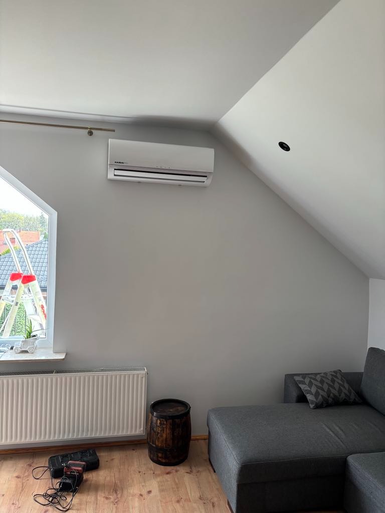 Zamontowana jednostka klimatyzacji na szarej ścianie poddasza, widoczna drabina oparta o okno, drewniana beczka, szara sofa i grzejnik.