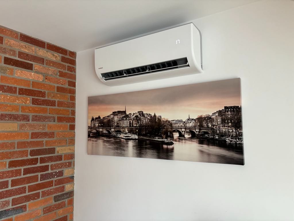 Biały klimatyzator ścienny marki Kaisai zamontowany nad czarno-białym obrazem miasta z rzeką, na białej ścianie obok ściany z czerwonej cegły.
