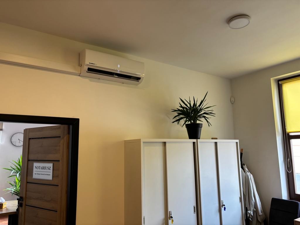 Biała klimatyzacja typu split zamontowana na ścianie w biurze notarialnym, widoczna nad wysoką białą szafą z przesuwnymi drzwiami i doniczką z zieloną rośliną na górze, w tle drzwi z tabliczką...