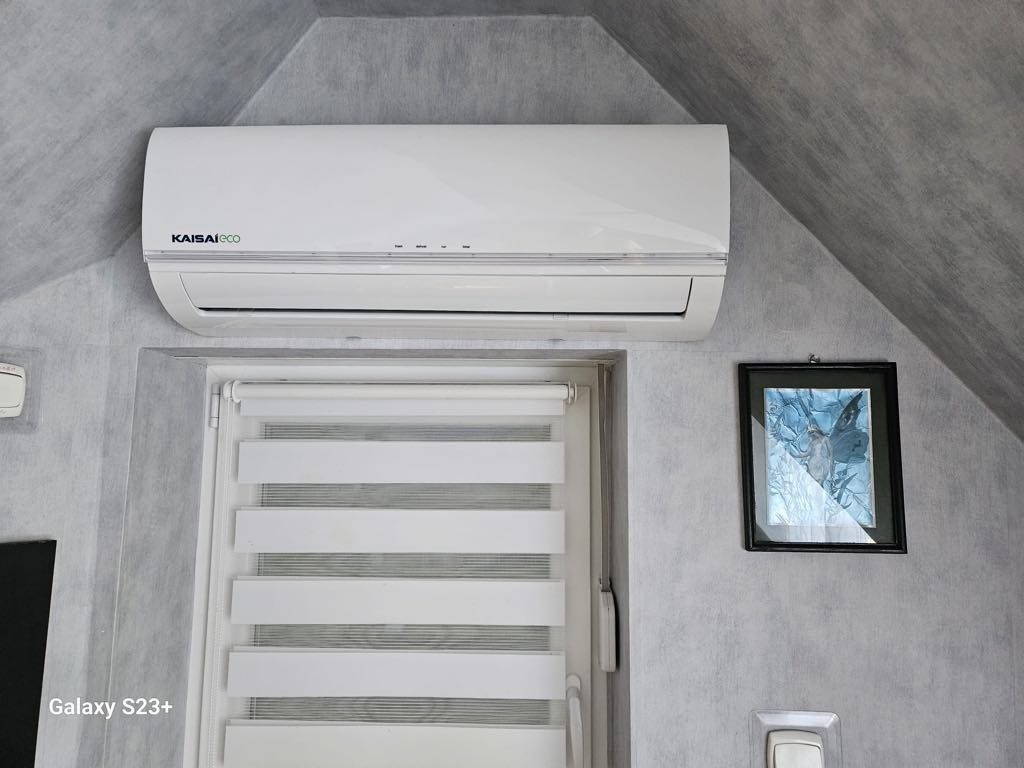 Biała jednostka klimatyzacyjna Kaisai Eco zamontowana na szarej ścianie pod skosem, nad oknem z żaluzjami i obok oprawionego obrazu.
