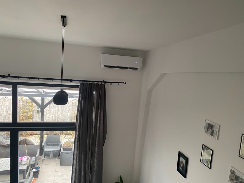 Biały klimatyzator ścienny w jasnym wnętrzu z dużym oknem i zasłoną, widok z góry, wisząca lampa kulista na tle okna.