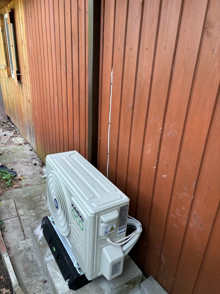 Zewnętrzna jednostka klimatyzacji Kaisai ECO, zamontowana na betonowym podeście przy drewnianej ścianie budynku z widocznym oknem w tle.
