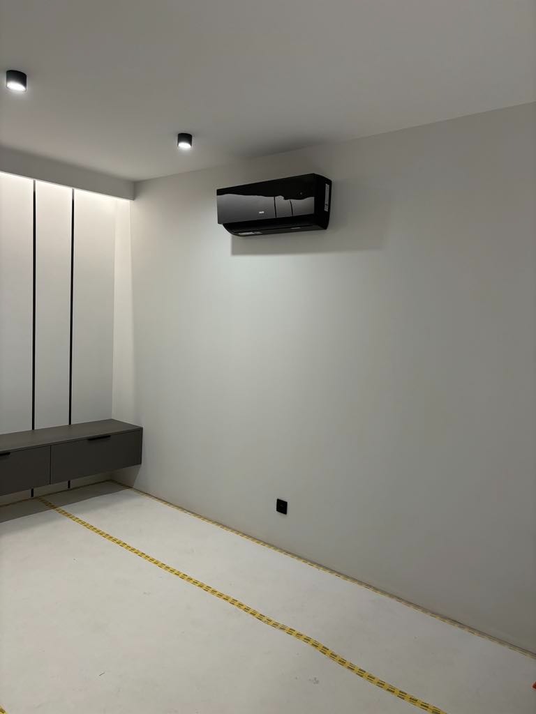 Czarny klimatyzator ścienny w minimalistycznym wnętrzu, widoczne taśmy malarskie na podłodze, szara szafka RTV i czarne punkty świetlne na suficie.