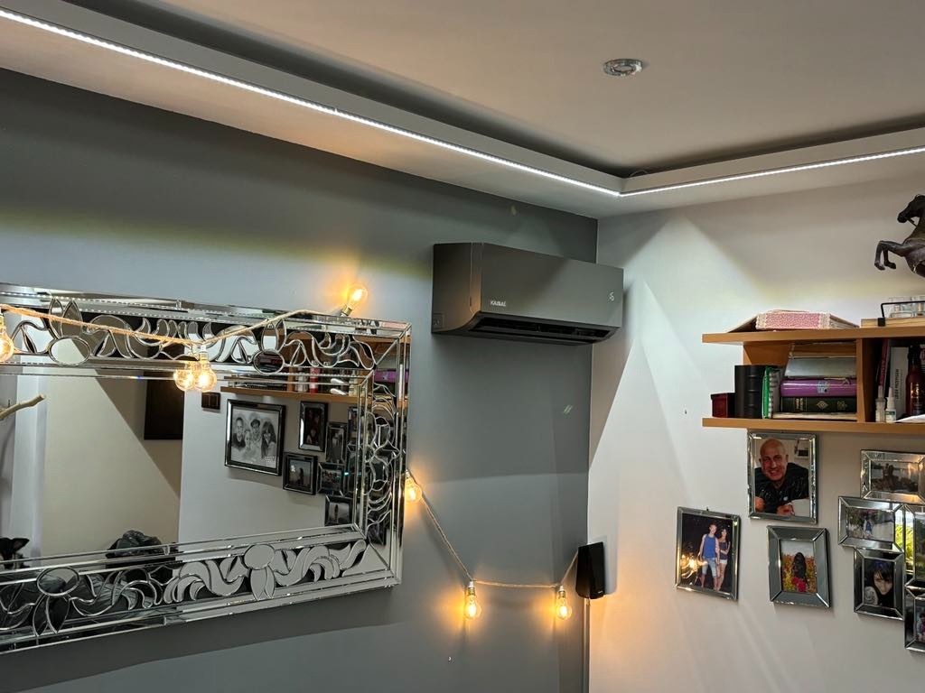 Szara klimatyzacja ścienna zamontowana w mieszkaniu w Lubinie, widoczna nad lustrem z ozdobną ramą i półką z książkami, oświetlona listwą LED na suficie.