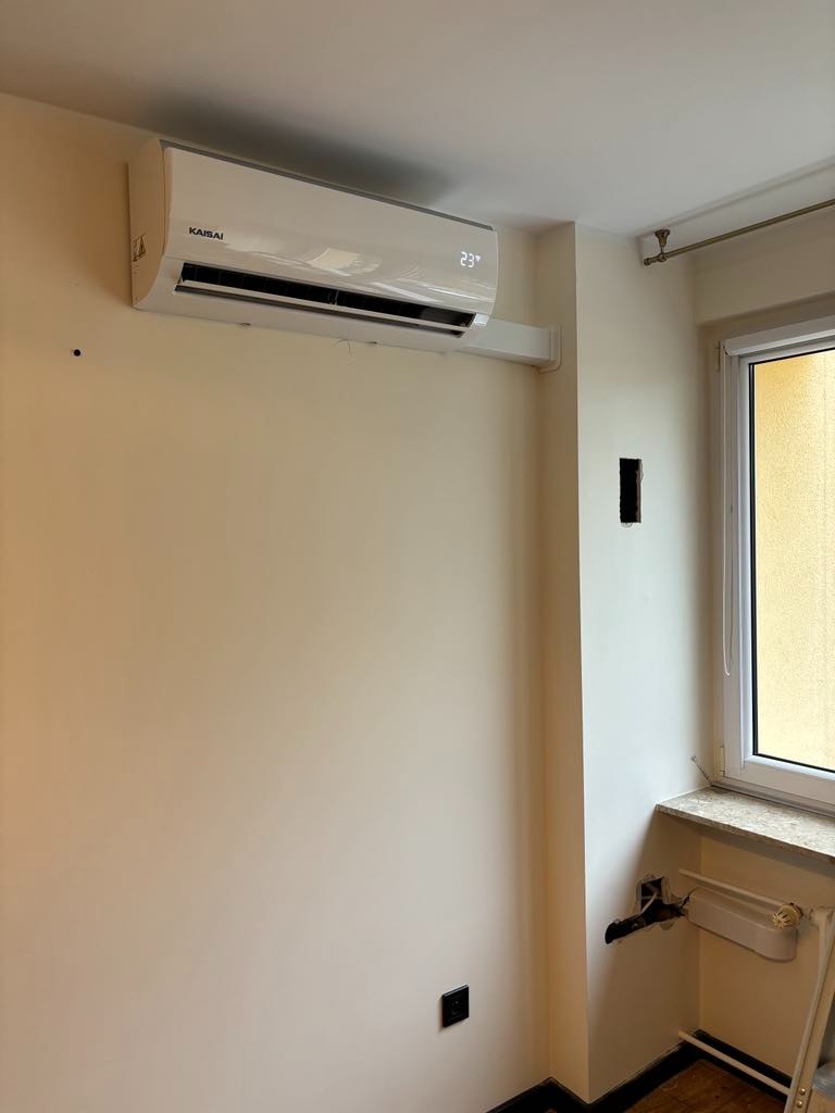 Biała jednostka klimatyzacyjna Kaisai zamontowana na kremowej ścianie w pomieszczeniu, widoczny wyświetlacz temperatury 23 stopnie Celsjusza, fragment okna i instalacja grzewcza w tle.