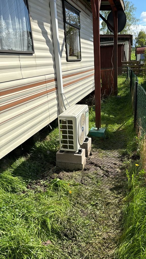 Zewnętrzna jednostka klimatyzacji zamontowana na betonowych bloczkach przy ścianie domku letniskowego, widoczna rynna odprowadzająca wodę, trawa wokół urządzenia.
