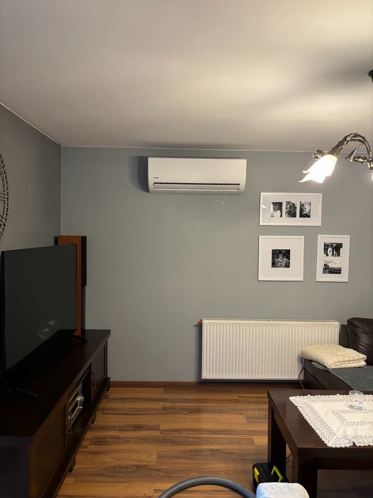 Biały klimatyzator ścienny zamontowany w szarym salonie nad kaloryferem, z ramkami ze zdjęciami na ścianie i elementami odkurzacza widocznymi w dolnej części kadru.