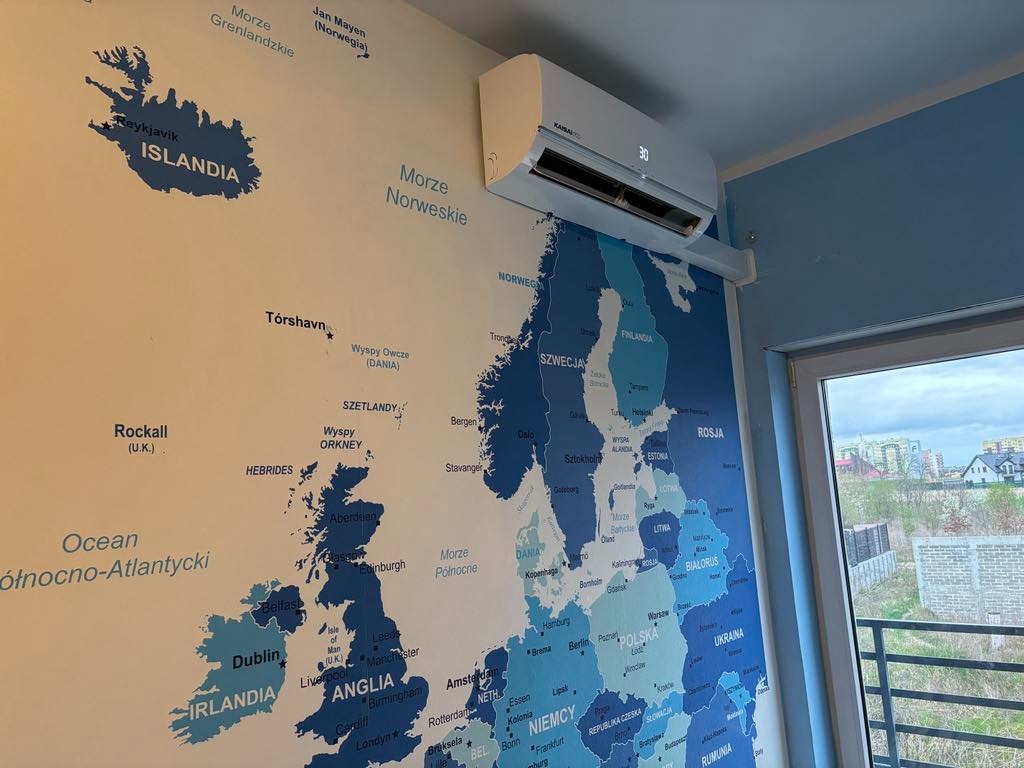 Biały klimatyzator ścienny marki Kaisai zamontowany w pokoju z tapetą przedstawiającą mapę Europy, widok z okna na osiedle domów.