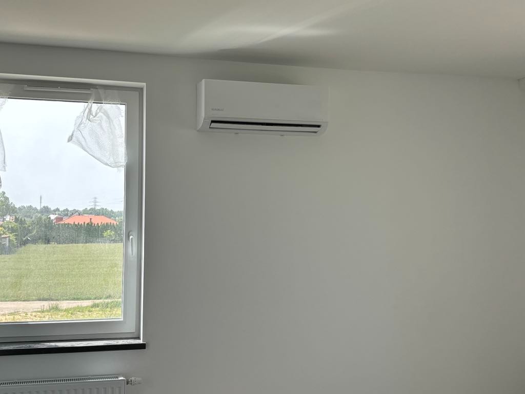 Biała jednostka klimatyzacji marki Kaisai zamontowana na białej ścianie w jasnym pomieszczeniu, widok przez okno na zielony trawnik i zabudowania.