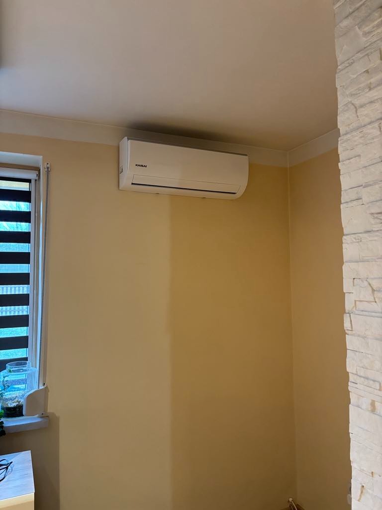 Biały klimatyzator marki Kaisai zamontowany na żółtej ścianie w pokoju z oknem i roletami, widoczny fragment ściany wykończonej kamieniem dekoracyjnym.