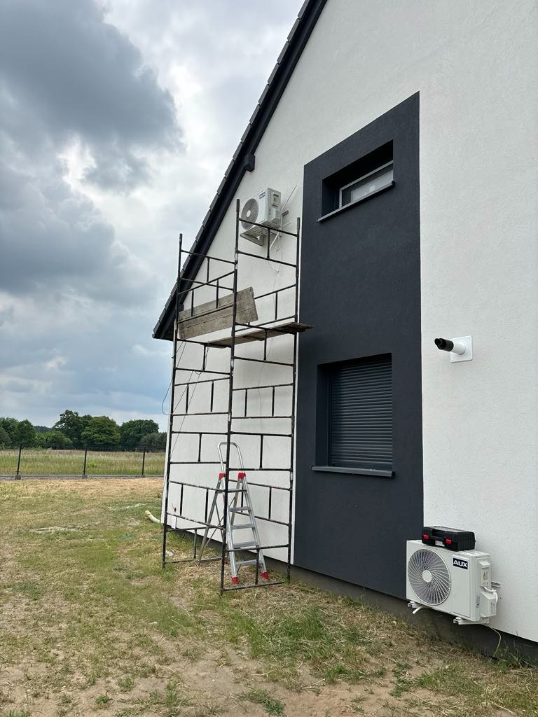 Montaż klimatyzacji zewnętrznej AUX na elewacji domu jednorodzinnego z użyciem rusztowania i drabiny, widok z boku na tle pochmurnego nieba.