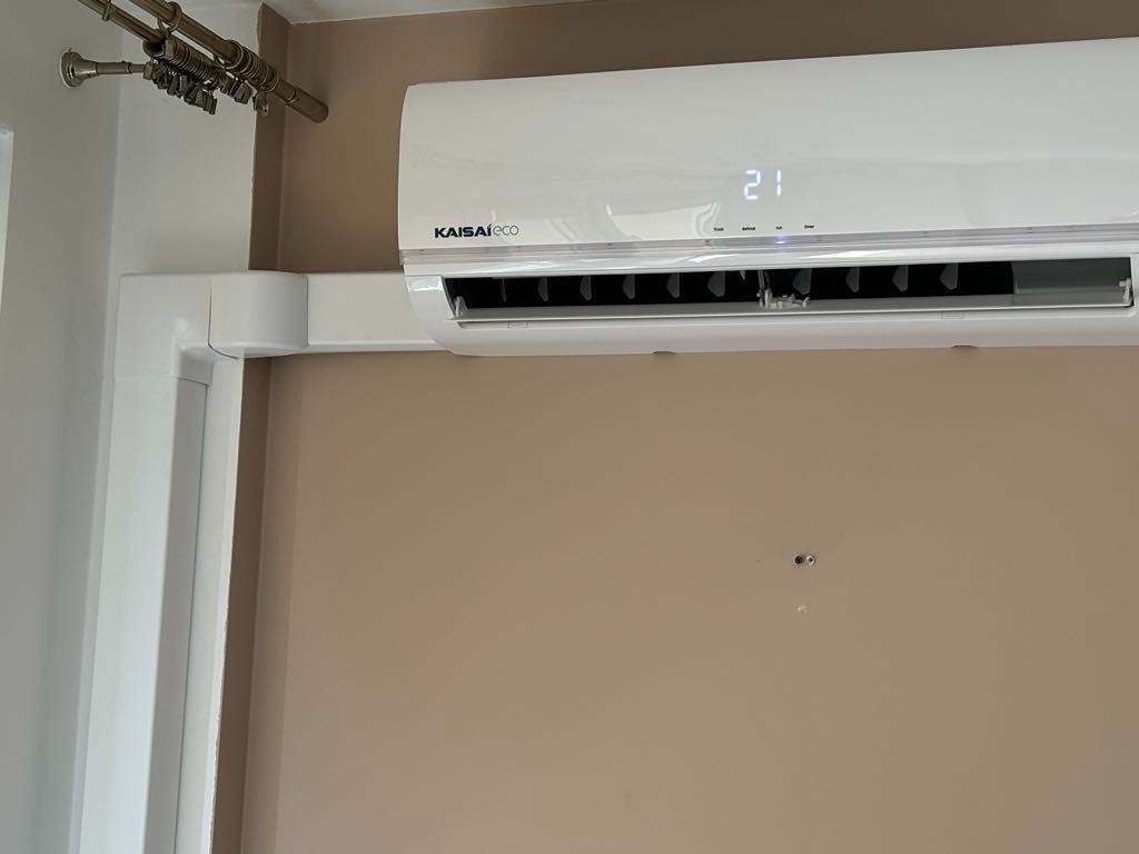 Biała jednostka klimatyzacji Kaisa Eco zamontowana na beżowej ścianie, widoczny wyświetlacz z temperaturą 21 stopni Celsjusza i fragment instalacji nad urządzeniem.