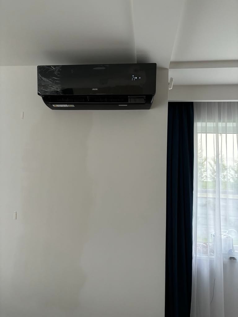 Czarny klimatyzator marki AUX zamontowany na białej ścianie w pokoju z zasłoniętym oknem.