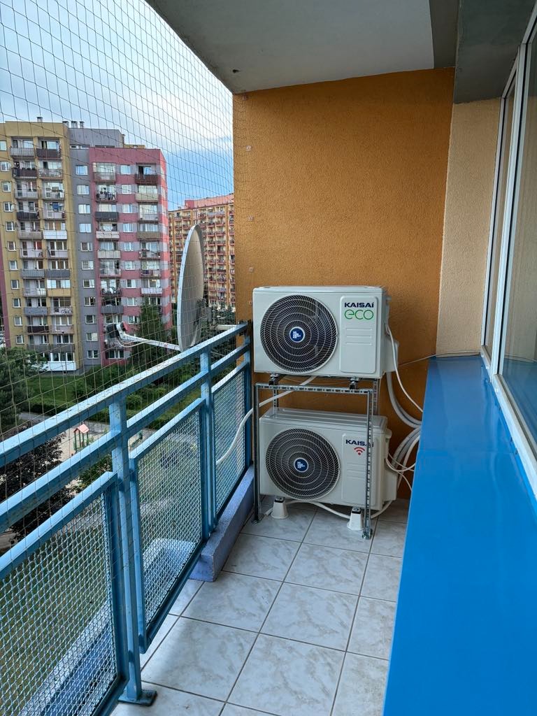 Dwa klimatyzatory Kaisai Eco zamontowane jeden nad drugim na balkonie mieszkania w bloku, widok na osiedle przez siatkę zabezpieczającą.