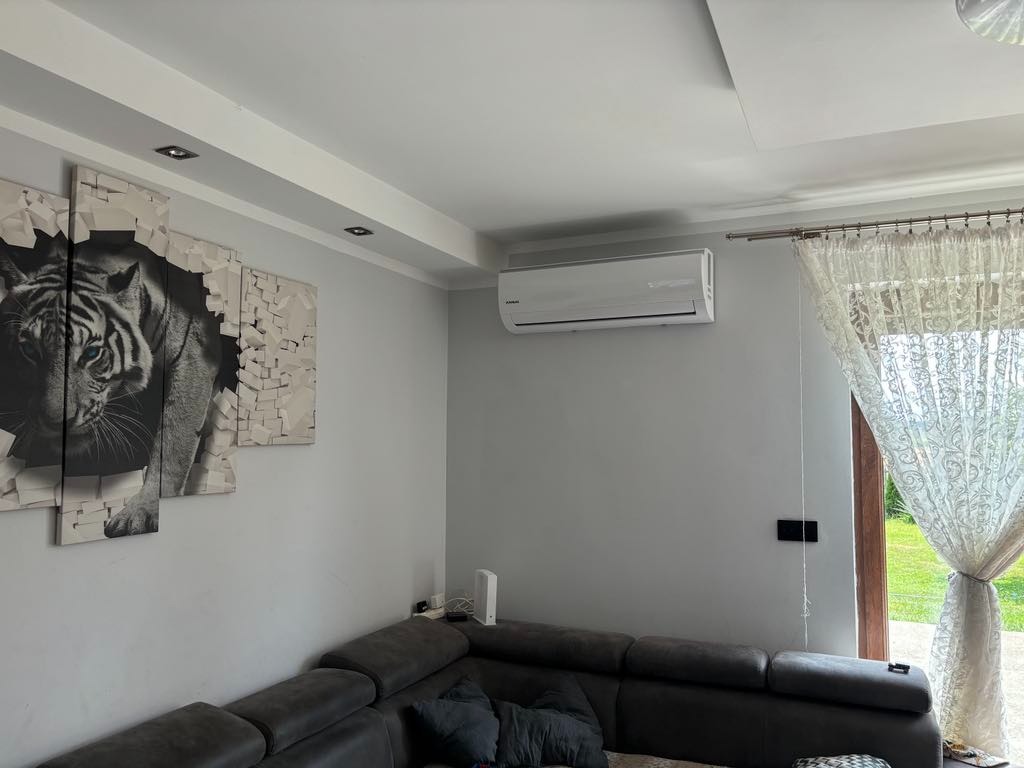 Biały klimatyzator na szarej ścianie w salonie z szarą sofą, obrazem tygrysa i firankami w oknie.